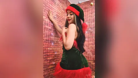JessieLiu poses against a brick wall wearing a short green velvet elf costume with red trim and a matching hat.