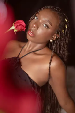 Beautiful Black woman with long braids in a black dress holding a red rose in a romantic, moody setting.