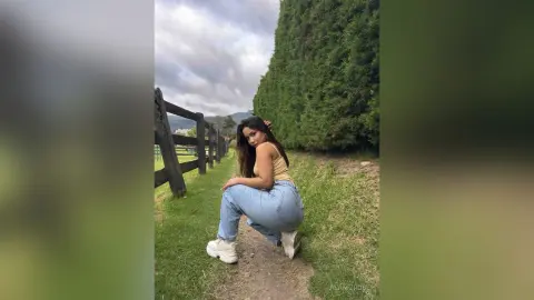 Young woman in a tan tank top and blue jeans sitting on a grassy path by a wooden fence under a cloudy sky.