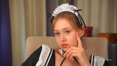 Melancholic young woman in a black and white maid outfit and frilled headband sitting in a room.