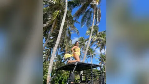 Cheerful woman in a yellow summer outfit sitting on a golf cart under tropical palm trees on a sunny day.