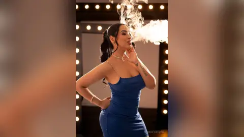 Glamorous woman in a blue sequin dress exhaling smoke in a studio with warm vanity lights.