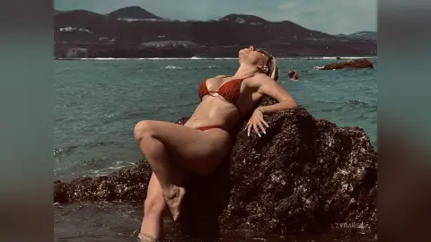 Blonde woman in a red bikini relaxing on coastal rocks by the ocean under a sunny sky.