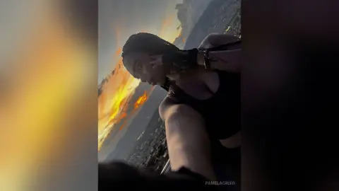 PamelaCreed wearing a black tank top, posing outdoors against a bright sunset or fire-lit sky.