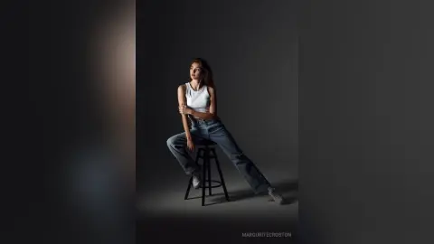 Young woman in white tank top and blue jeans sitting on a stool in a moody studio setting.