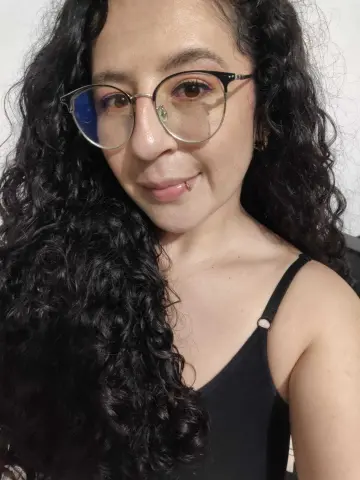 Smiling woman with long curly black hair and round glasses wearing a black tank top in a bright indoor setting.