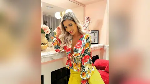 Smiling blonde woman in a colorful floral blouse posing in a bright, modern room.