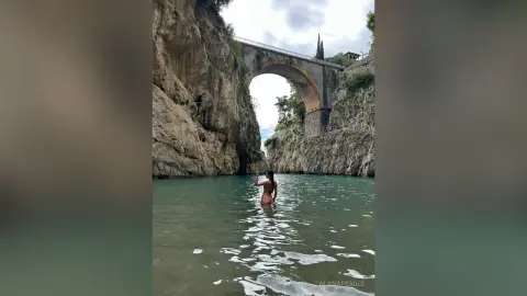 Woman in a bikini wading in turquoise river water beneath a massive stone arch bridge in a rocky canyon.