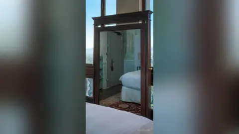Modern bedroom interior featuring a large dark wood framed mirror reflecting a bright, airy room with white bedding.