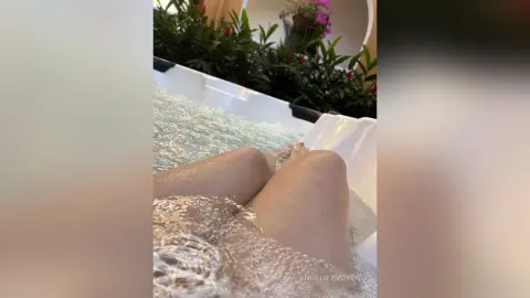 Relaxed woman soaking in a bubbly outdoor jacuzzi surrounded by lush green plants and summer sunlight.