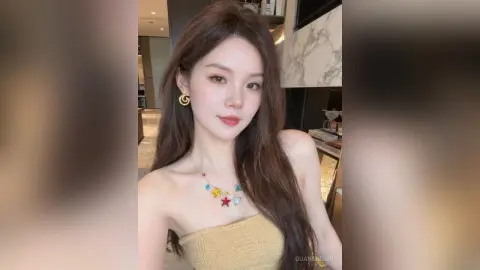 Young woman with long brown hair wearing a gold strapless top and colorful star necklace in a modern interior.