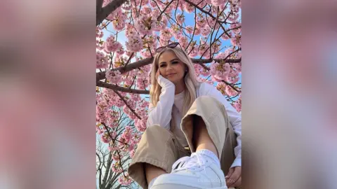 Blonde woman in white long-sleeve shirt and khaki pants relaxing under blooming pink cherry blossom trees.