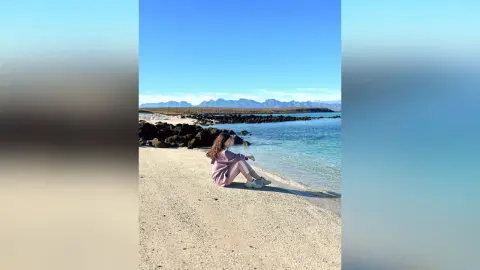 KiraManila — Woman in a pink sweater sitting on a sandy beach by turquoise ocean water under a clear blue sky.