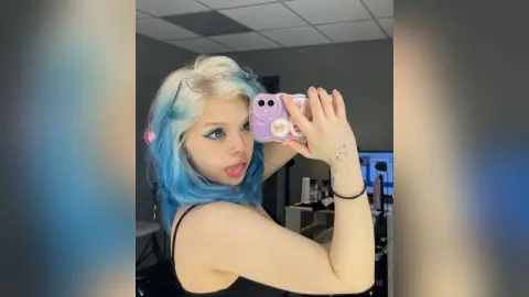 Young woman with blue and blonde hair taking a playful mirror selfie in a black tank top.