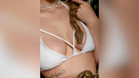 Woman in white bikini top holding a large brown snake in a natural outdoor setting.