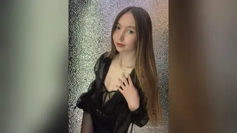 Young woman in a black sheer blouse posing against a shimmering silver glitter background.