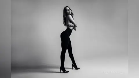 Elegant woman in black leggings and high heels posing in a minimalist black and white studio setting.