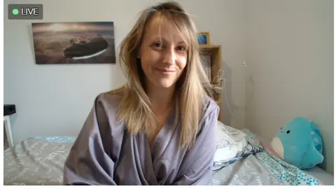Smiling blonde woman in a grey silk robe relaxing on a bed in a bright bedroom.