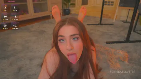 Playful young woman with long brown hair sticking her tongue out while lying on a grey rug in a cozy room.