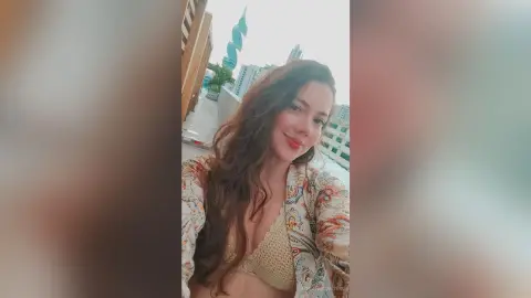 Smiling brunette woman in a patterned bohemian cardigan and bikini top on a sunny urban rooftop.
