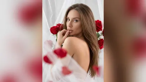 Beautiful woman in a white sheer outfit surrounded by red roses in a dreamy, romantic studio setting.