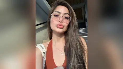 Young woman with long hair wearing a rust orange tank top and glasses in a bright indoor setting.