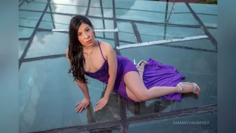 SamanthaHarvey poses on a glass roof wearing a purple slip dress with a high slit.