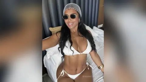 Glamorous woman in white bikini and sunglasses relaxing on a bed in a bright, chic bedroom setting.