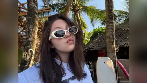Young woman in white sunglasses and t-shirt relaxing under palm trees at a tropical beach resort.