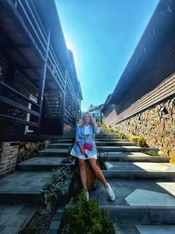 NikaZen sits on outdoor stone steps wearing a short blue dress and white sneakers under a bright blue sky.