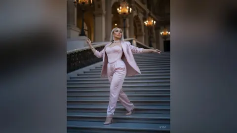 Elegant woman in a blush pink satin pantsuit posing on grand marble stairs in a luxury classical building.
