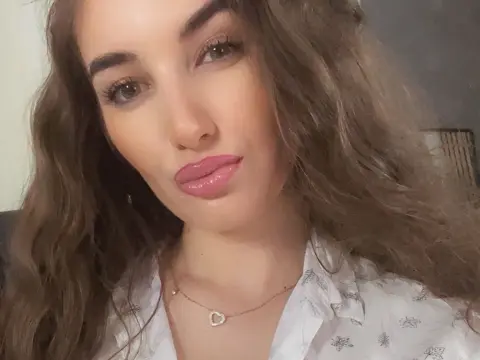 Close-up portrait of a young woman with wavy brown hair wearing a white patterned blouse in soft indoor lighting.