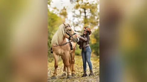 Smiling female equestrian in jeans and boots bonding with a blonde horse in a sunny autumn forest.