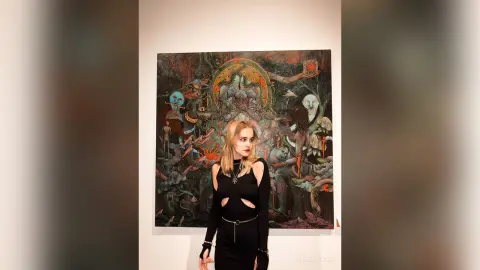 Young woman in a black cutout dress standing before a surreal fantasy landscape painting in an art gallery.