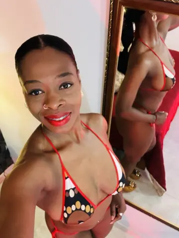 Smiling Black woman in a red and white patterned bikini posing for a mirror selfie in a bright room.