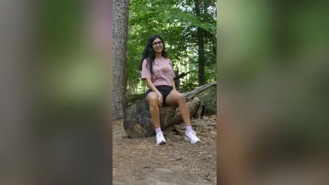 Smiling young woman in a pink graphic t-shirt and black shorts sitting on a log in a lush green forest.