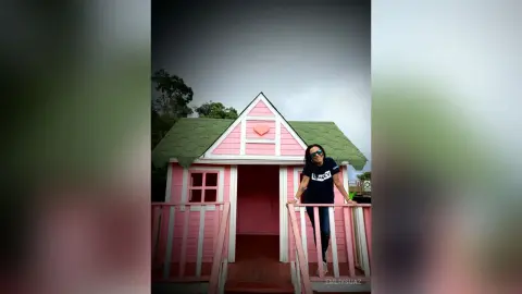 Smiling woman in a navy t-shirt standing in front of a whimsical pink wooden playhouse under a cloudy sky.