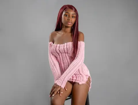 Young woman with long red hair wearing a pink off-the-shoulder knit sweater sitting against a grey background.