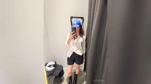 Young woman in a white button-down shirt, black shorts, and blue baseball cap taking a mirror selfie in a room.
