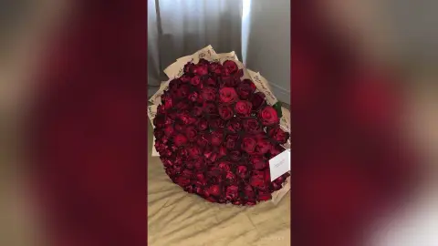 Large romantic bouquet of deep red roses in brown wrapping paper on a neutral surface.
