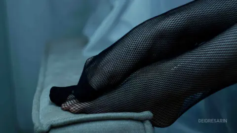 Close-up of legs in black fishnet tights resting on a soft white textured surface in moody blue lighting.