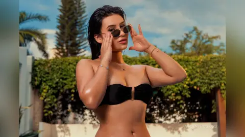 Stylish woman in black bikini top and sunglasses posing outdoors against a lush green garden background.