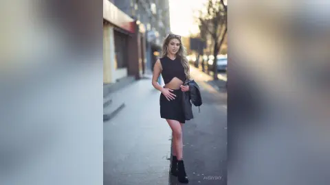 Stylish young woman in a black crop top and mini skirt standing on a city sidewalk during golden hour.