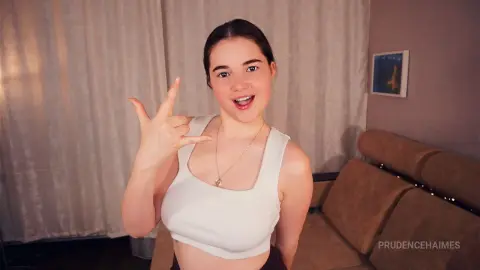 Smiling young woman in a white crop top making a hand gesture in a cozy living room setting.