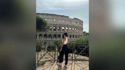 Woman in white tank top and black trousers standing before the historic Roman Colosseum on a sunny day.