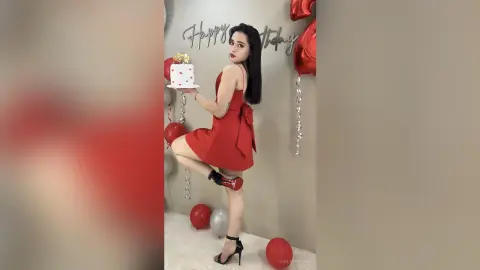 Young woman in a red cocktail dress holding a birthday cake near red balloons in a festive room.
