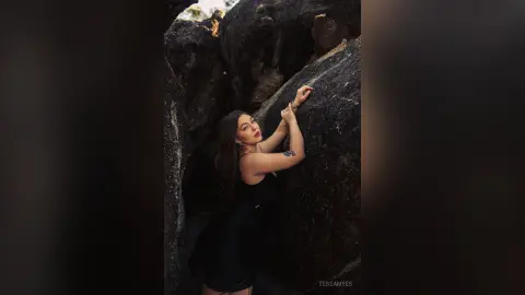 Woman in a black dress leaning against dark rock formations in a moody, cinematic outdoor setting.
