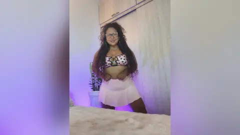 Smiling woman with curly hair in a polka dot bikini top and pink skirt posing in a bright room.