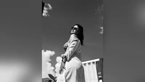 JulianaNavarro poses outdoors in a white robe and sunglasses, looking back over her shoulder against a cloudy sky.