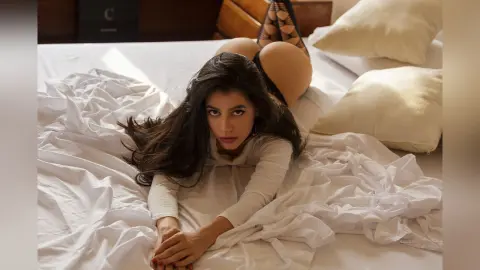 JessieLiu lies prone on white bedsheets, wearing a white long-sleeved top that exposes her bare buttocks.
